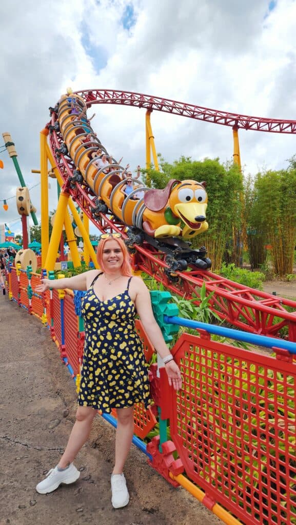A woman poses in front of a roller coaster in the shape of slinky dog from toy story.