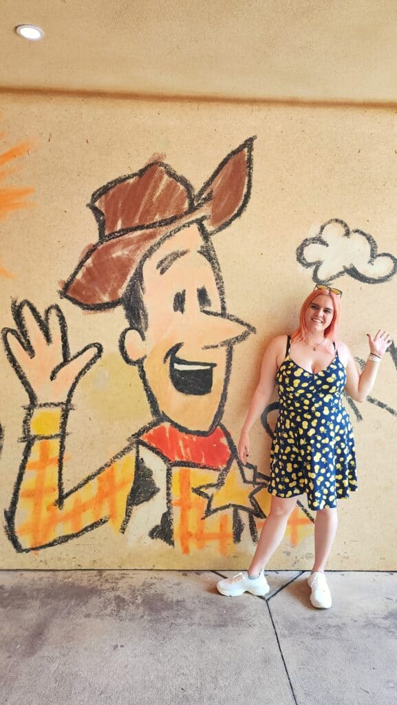 A woman waves in front of a wall showing a mural of Woody from Toy Story waving.