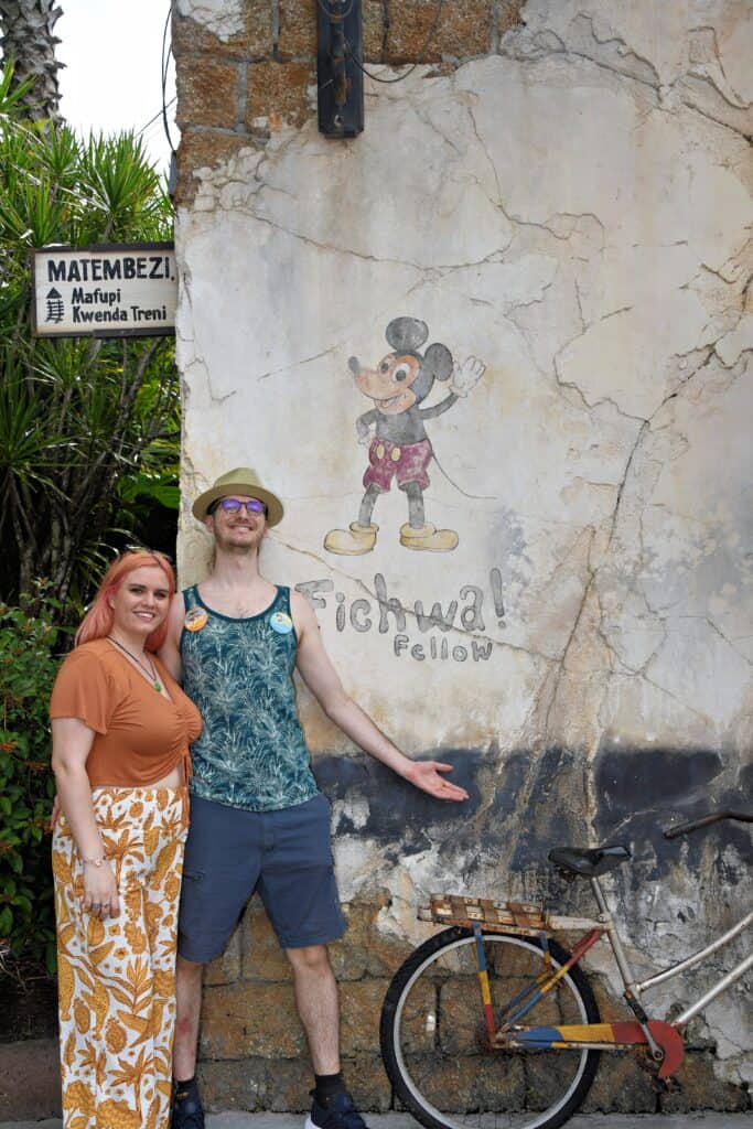 A man and woman stand in front of a mural on the wall showing Mickey Mouse and the words "Fichwa! Fellow."