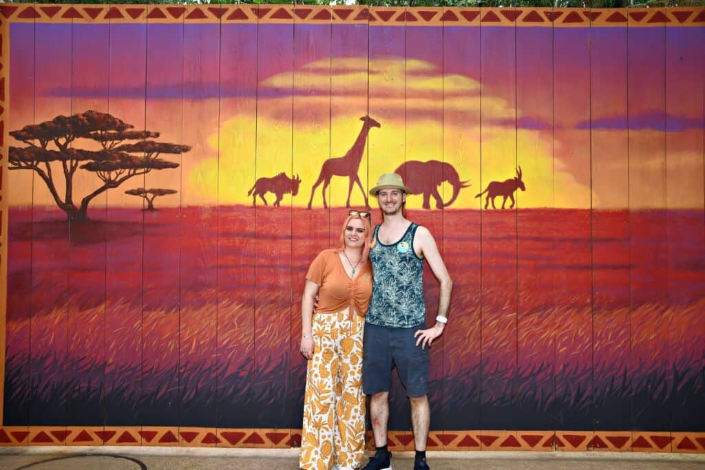 A woman and man stand in front of a bright and colorful "Lion King"-themed wall.