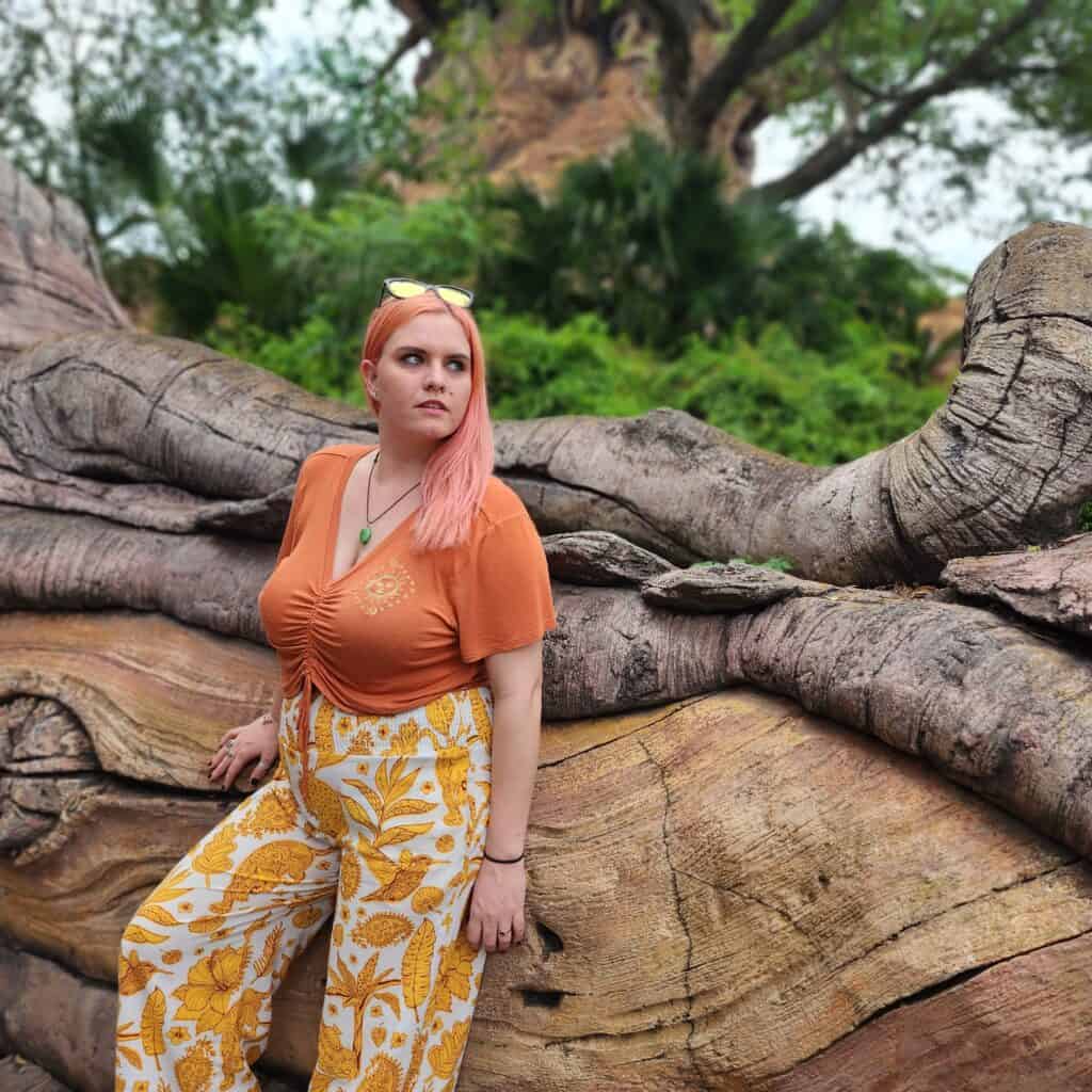 A woman leans against very large tree roots in a jungle setting.