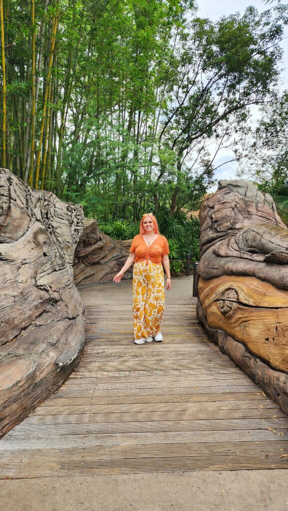A woman stands on a wooden bridge next to very large tree roots.