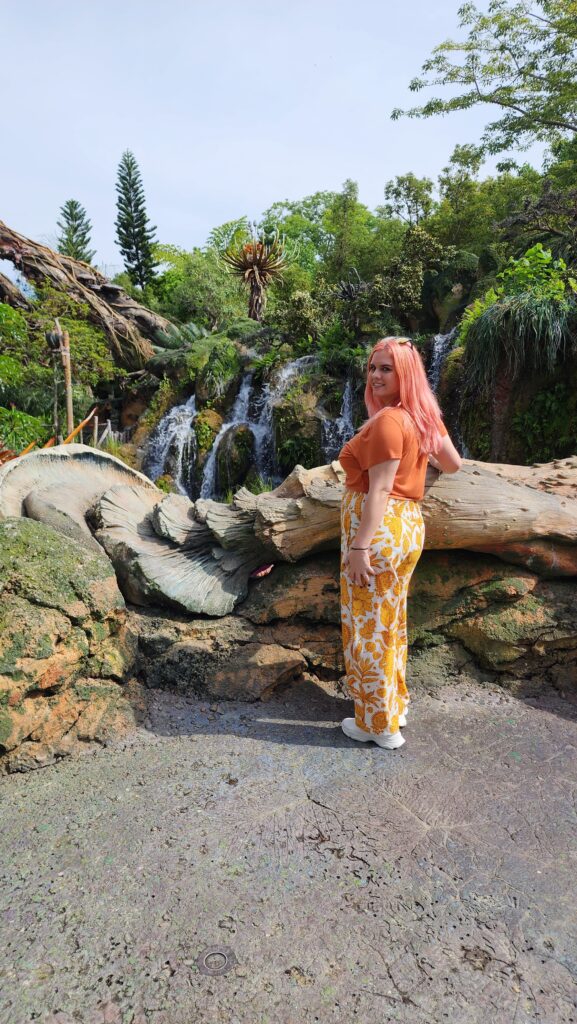 A woman stands in front of a waterfall in a jungle setting.