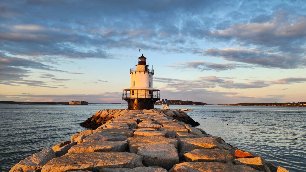 A lighthouse at the end of a rocky pathway next to the ocean at sunset.