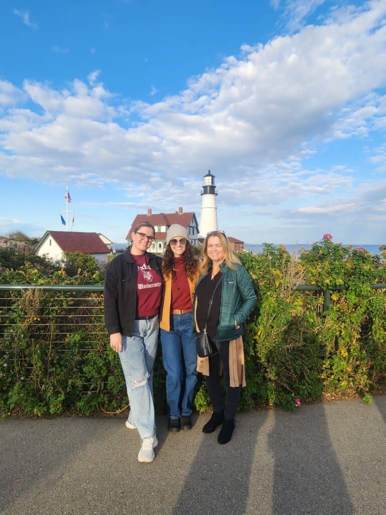 Three women stand in front of a lighthouse in Portland, Maine
