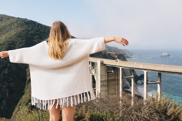 A woman in an oversized poncho stands with her arms outstretched like wings, overlooking a bridge built near the ocean coast. She's probably contemplating about slow travel. 