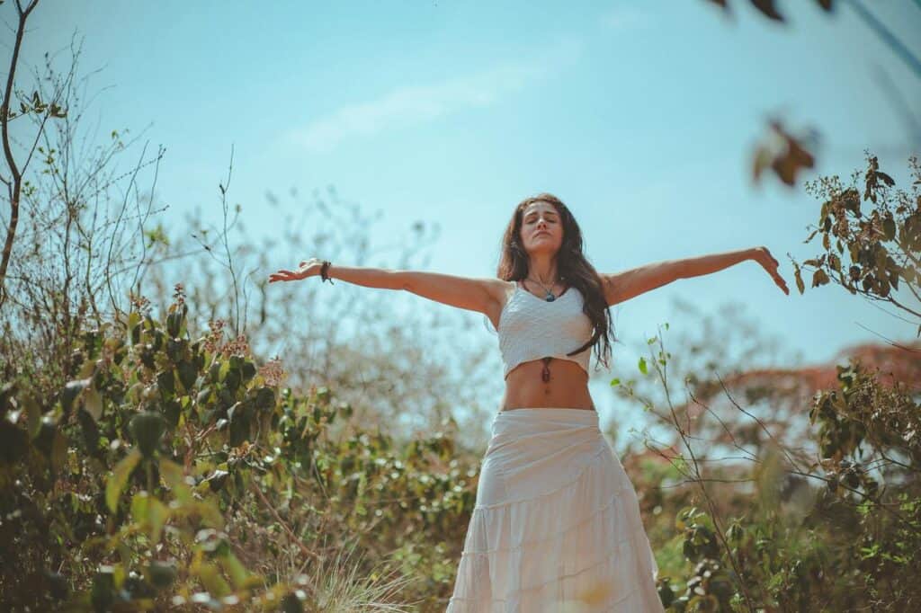 A woman stands in nature, eyes closed, arms spread out wide.