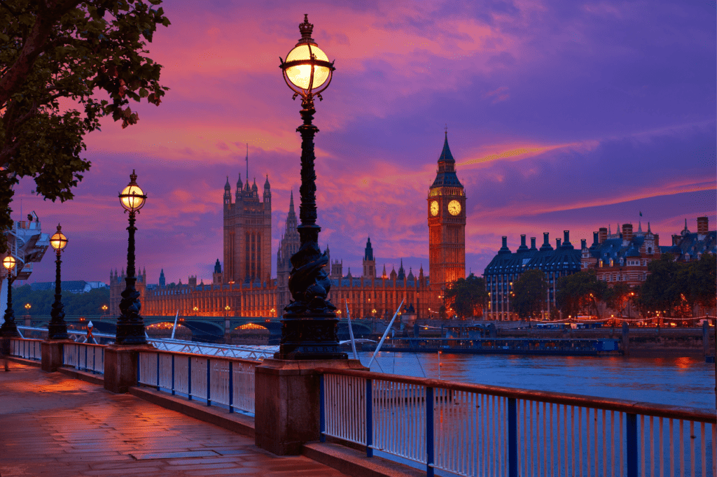 Street lamps light up a dusk sky; a clock tower sits on the other side of a river.
