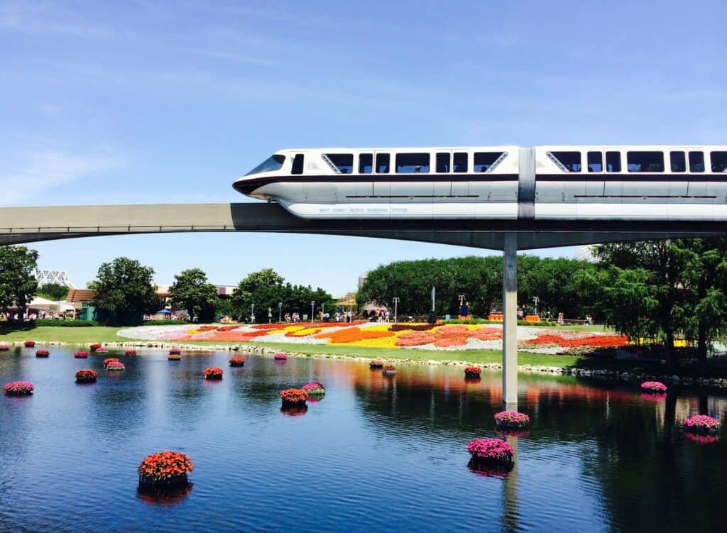 A monorail zooms past a body of water surrounded by colorful flowers. 