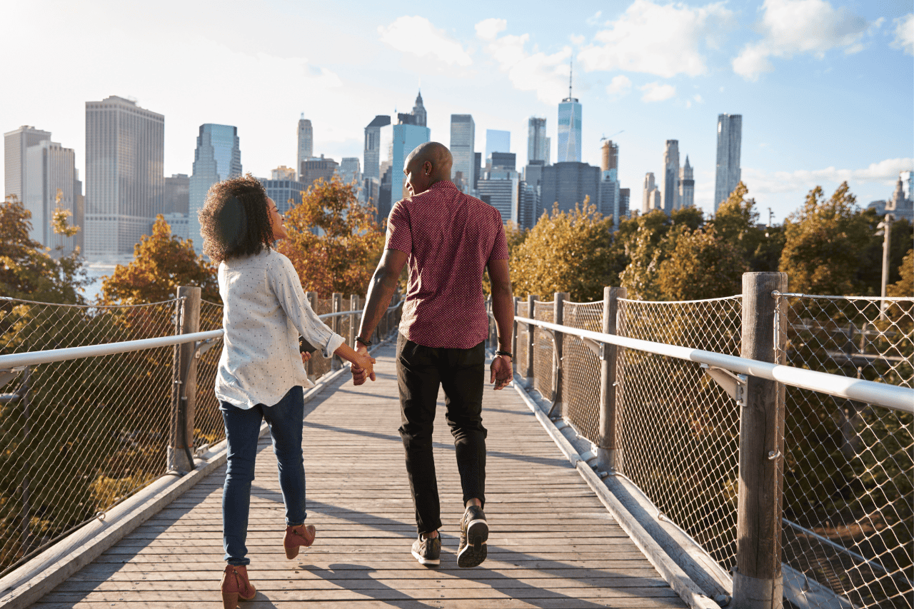 A man and woman holding hands cross a bridge in a park with the New York City skyline in the distance.