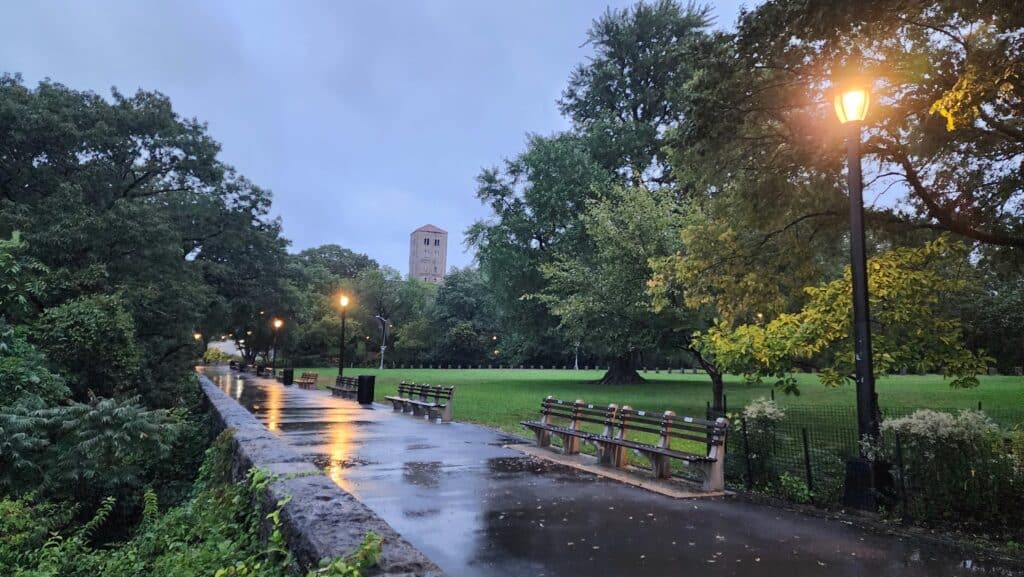 A light post illuminates the walkway in a park. A tower peers over the trees in the background.