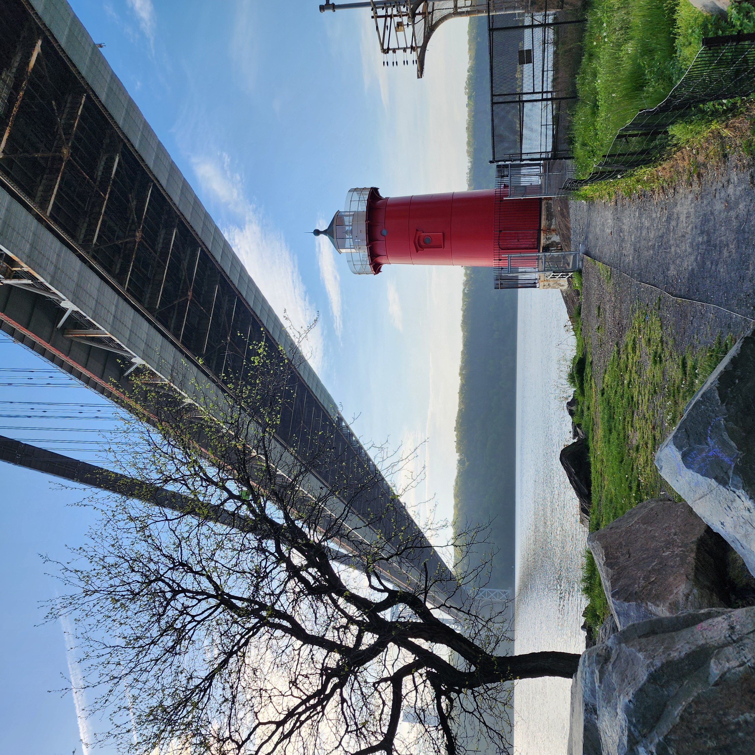 A little red light house stands under a bridge crossing over a large river on a clear day.